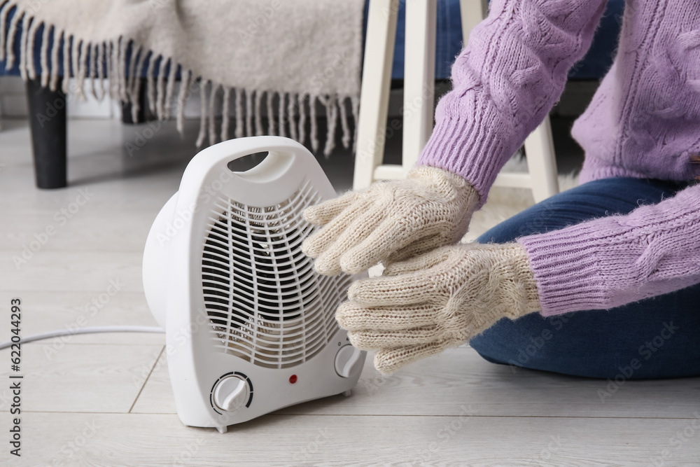 Woman warming hands in gloves near electric fan heater at home, closeup