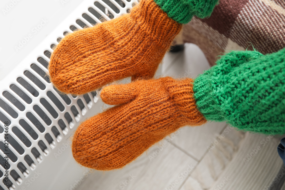 Woman warming hands in mittens near radiator at home, closeup