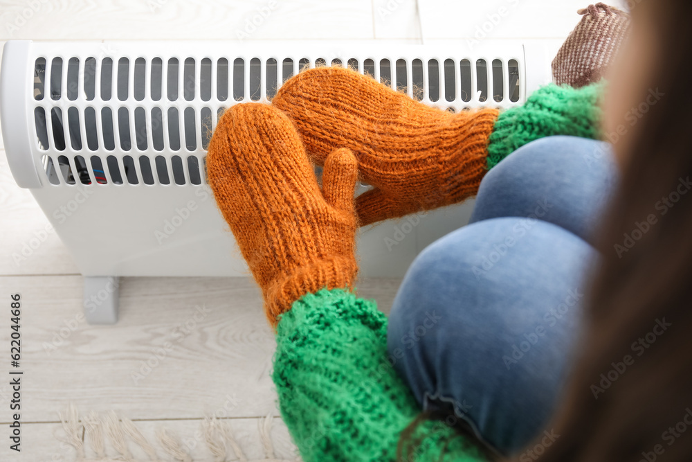 Woman warming hands in mittens near radiator at home, closeup