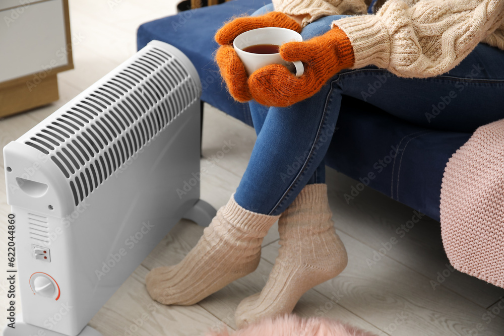 Woman with cup of hot coffee warming near radiator at home, closeup