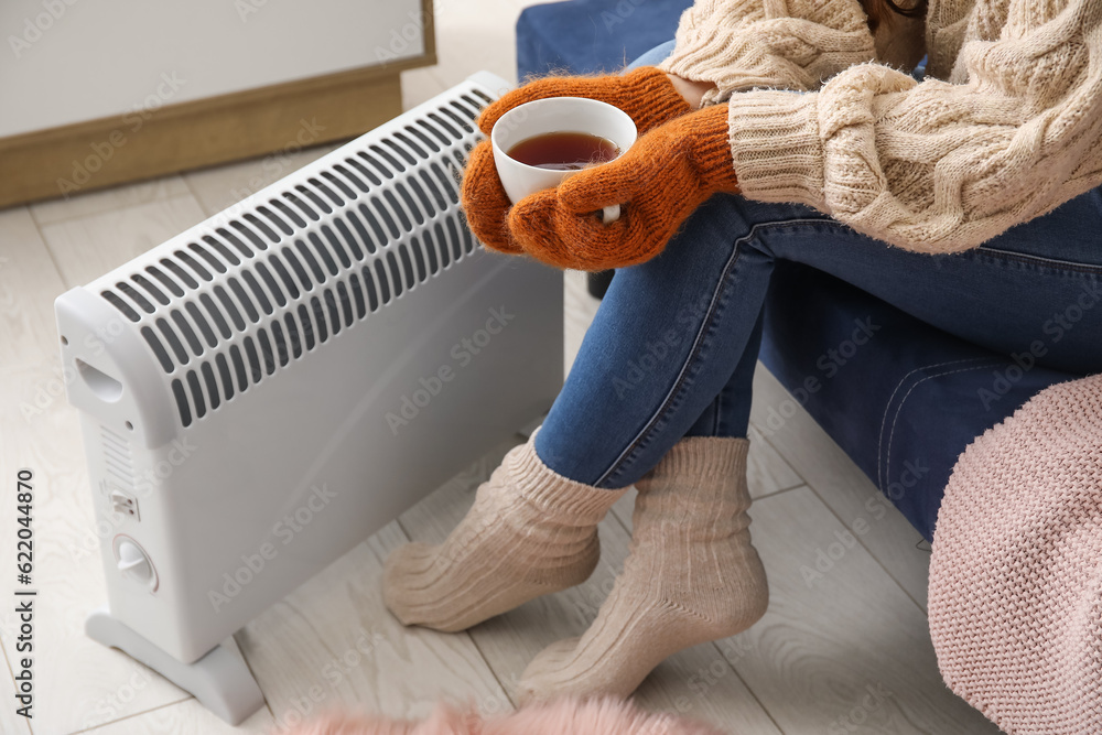 Woman with cup of hot coffee warming near radiator at home, closeup