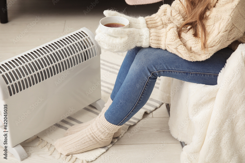 Woman with cup of hot coffee warming near radiator at home, closeup