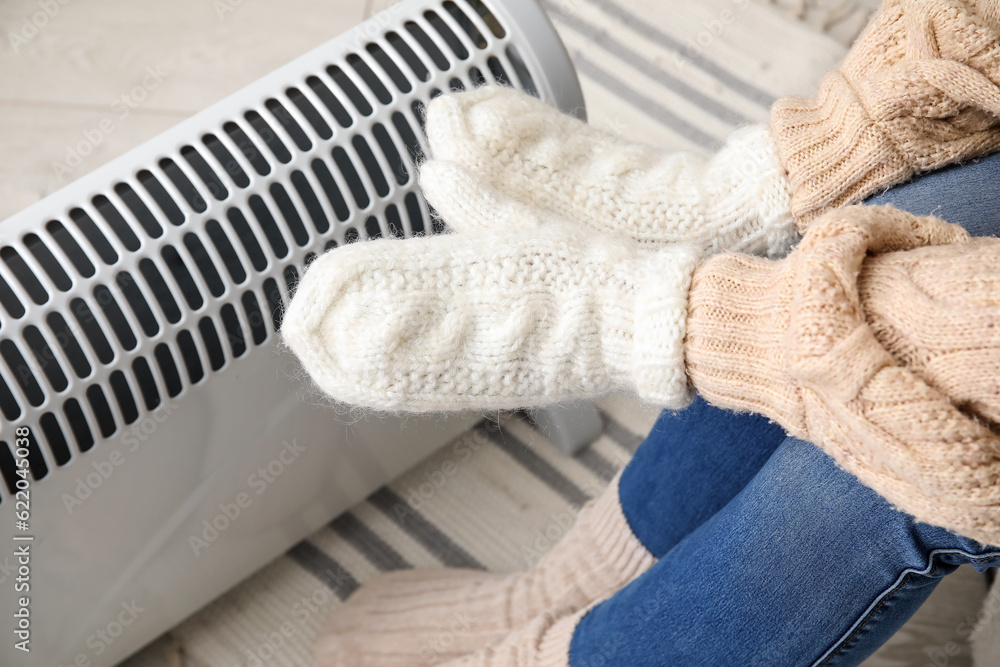 Woman warming hands in mittens near radiator at home, closeup