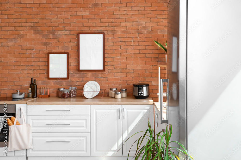 Interior of kitchen with stylish fridge, counters and blank pictures hanging on brick wall