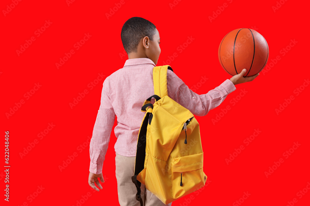 Little African-American schoolboy with ball and backpack on red background, back view