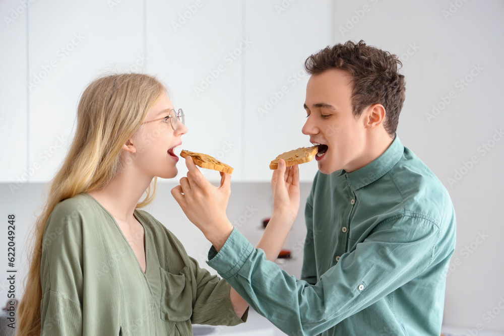 Young beautiful couple feeding each other tasty toasts of nut butter in kitchen