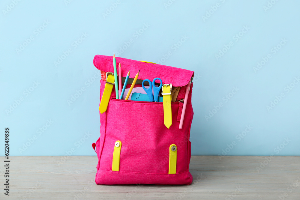 Pink school backpack with scissors, notebooks and pencils on grey wooden table near blue wall