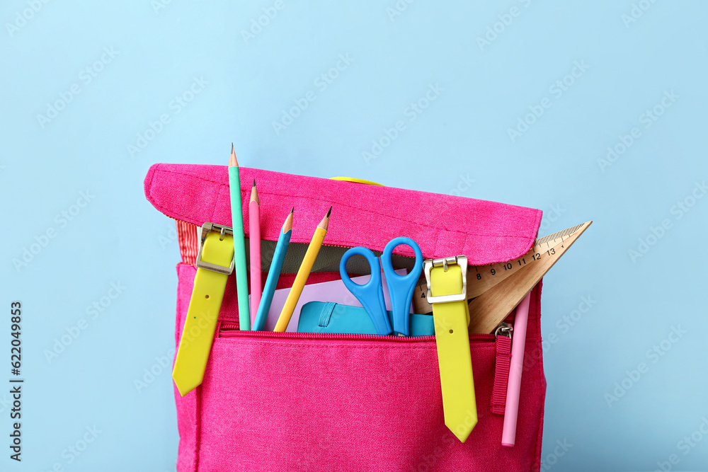 Pink school backpack with scissors, notebooks and pencils near blue wall