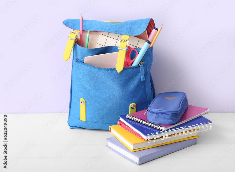 Blue school backpack with books, pencil case and markers on white wooden table near lilac wall