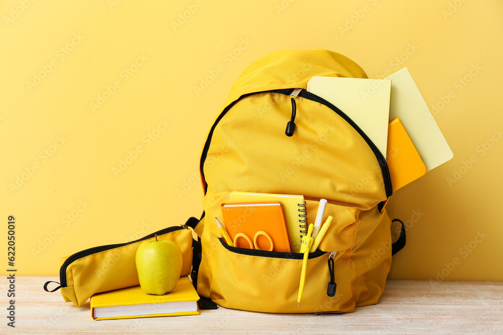 Yellow school backpack with notebooks, apple and scissors on wooden table near wall