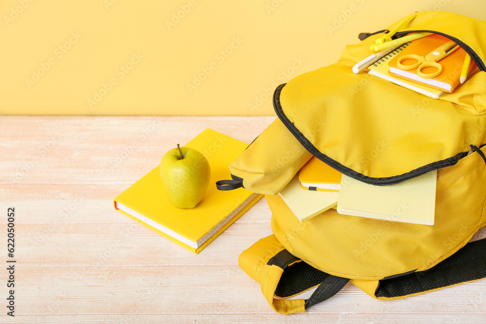 Yellow school backpack with notebooks, apple and scissors on wooden table near wall