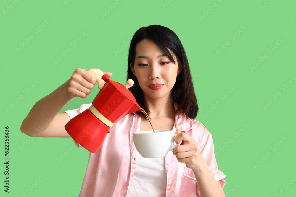 Beautiful Asian woman in pajamas pouring coffee into cup on green background