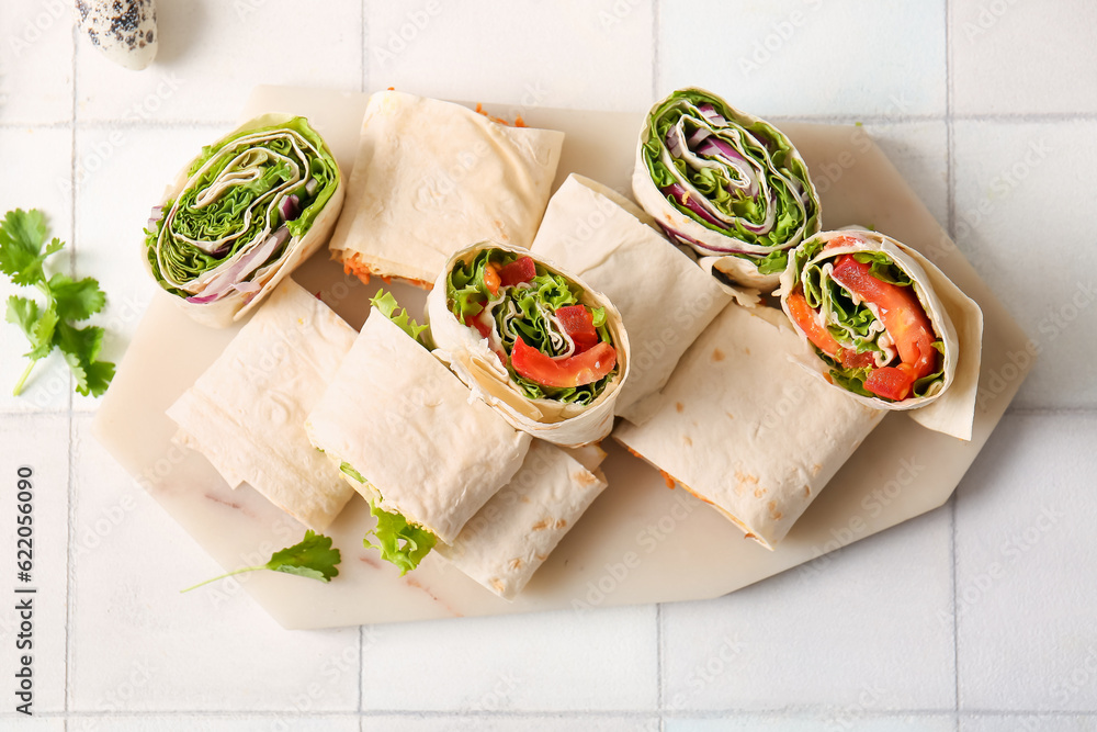 Board of tasty lavash rolls with vegetables on white tile background