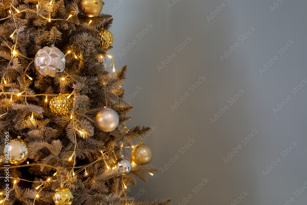 Christmas tree decorated with glowing lights, golden and silver balls near blue wall