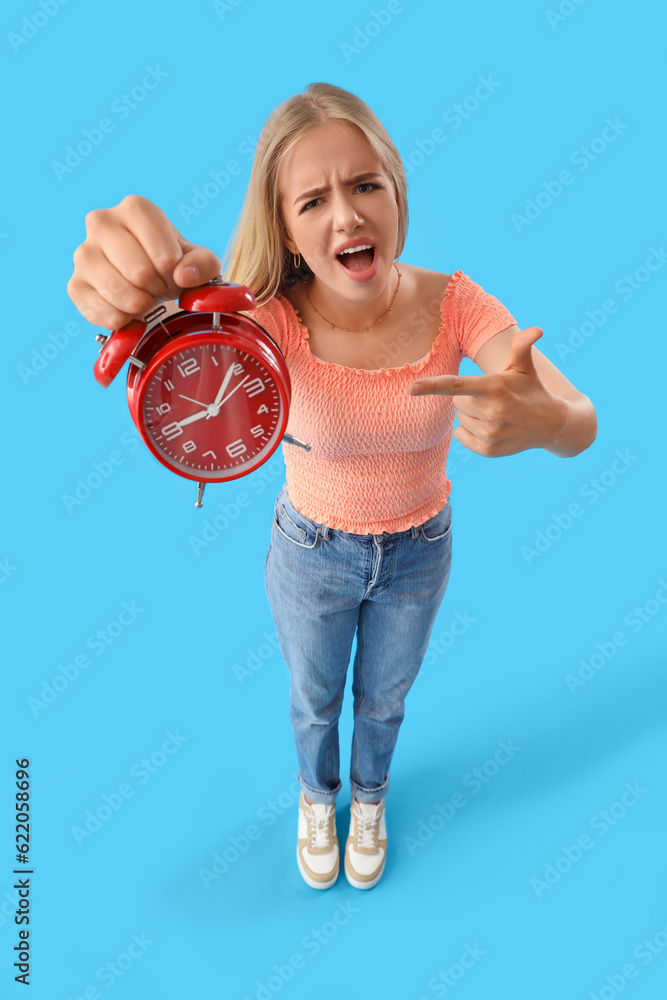 Stressed young woman pointing at alarm clock on blue background. Deadline concept