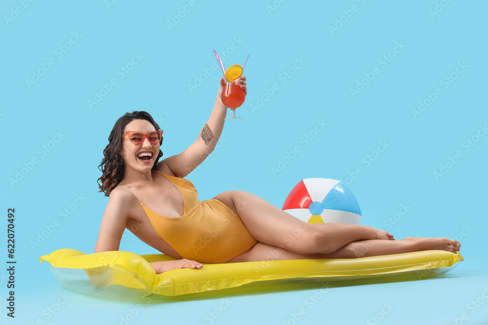 Young woman in swimsuit with cocktail and inflatable mattress on blue background