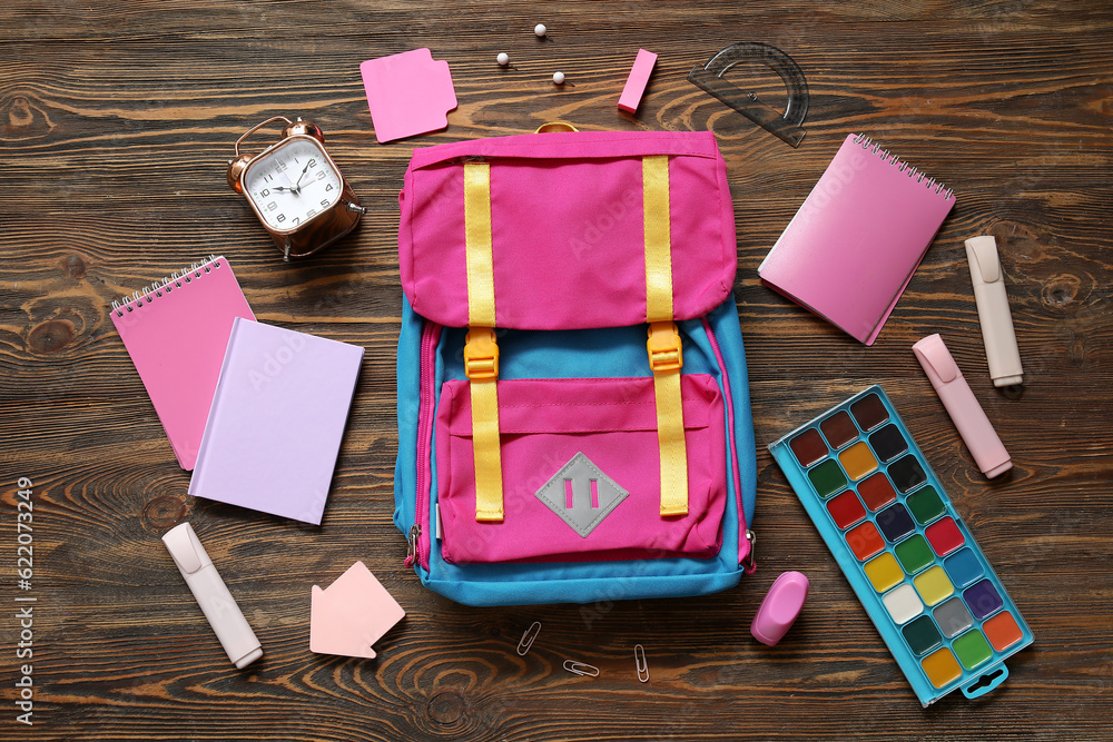 Colorful school backpack with notebooks, watercolors and alarm clock on brown wooden background