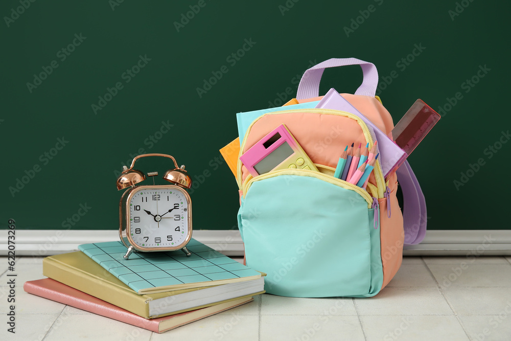 Colorful school backpack with notebooks, alarm clock and calculator on white tile table near green c