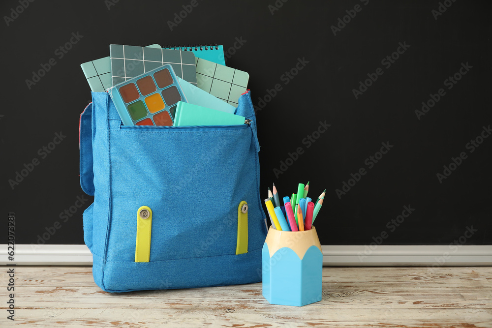 Blue school backpack with notebooks and cup of pencils and markers on wooden table near black chalkb