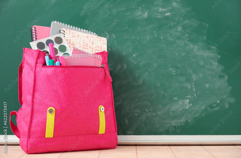 Pink school backpack with notebooks, watercolors and markers on beige tile table near green chalkboa