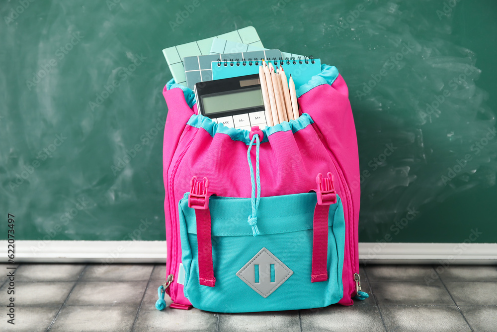 Colorful school backpack with notebooks, calculator and pencils on grey tile table near green chalkb