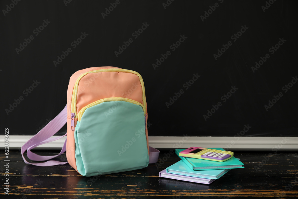 Colorful school backpack with notebooks and calculator on wooden table near black chalkboard