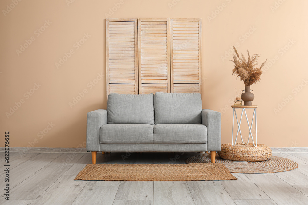 Stylish grey sofa, folding screen, table and pampas grass near beige wall in room