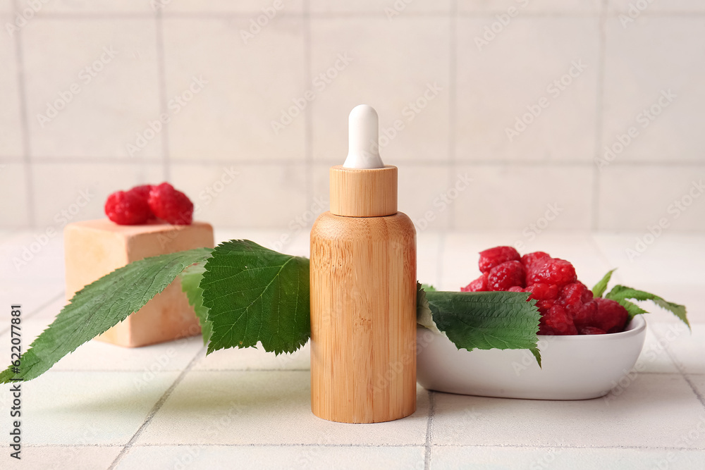 Bottle of cosmetic raspberry oil and bowl with berries on white tile background