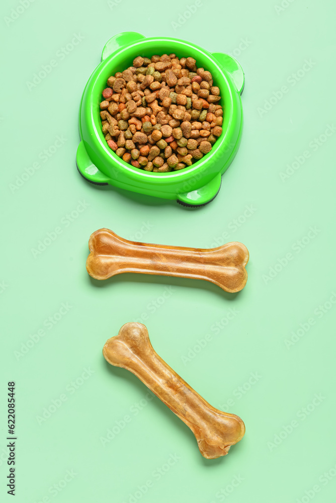 Bowl with dry dog food and chew bones on color background