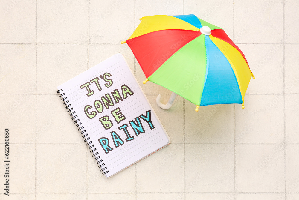 Notebook with text ITS GONNA BE RAINY and umbrella on white tile background. Weather forecast conce