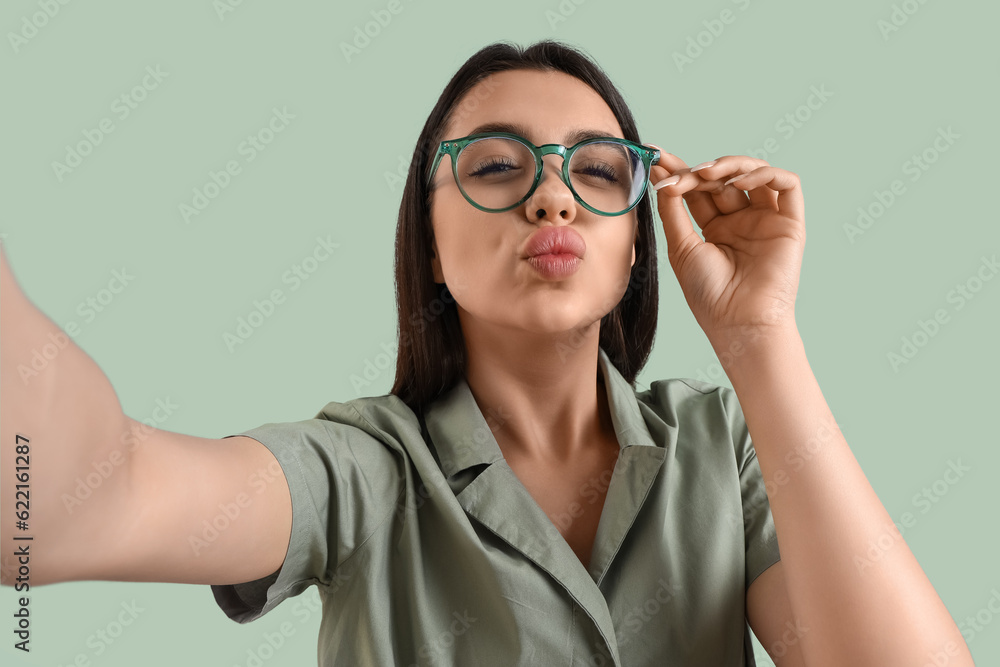 Beautiful young woman wearing glasses and taking selfie on color background