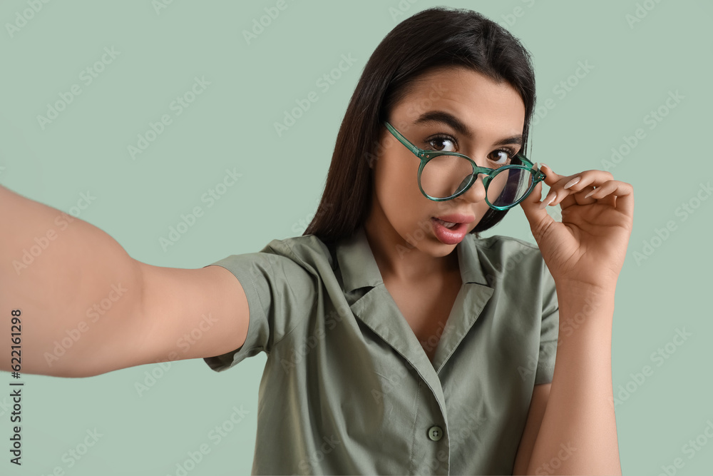 Beautiful young woman wearing glasses and taking selfie on color background