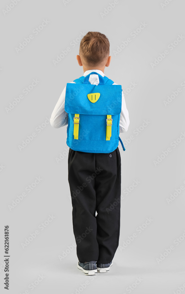 Little schoolboy with blue backpack on grey background, back view