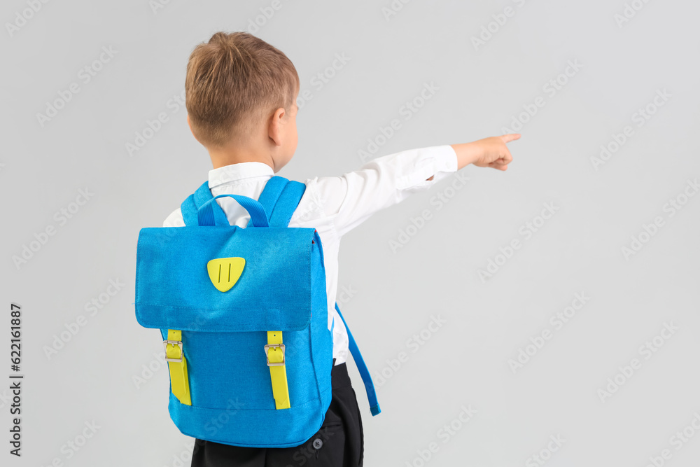 Little schoolboy with blue backpack pointing at something on grey background, back view