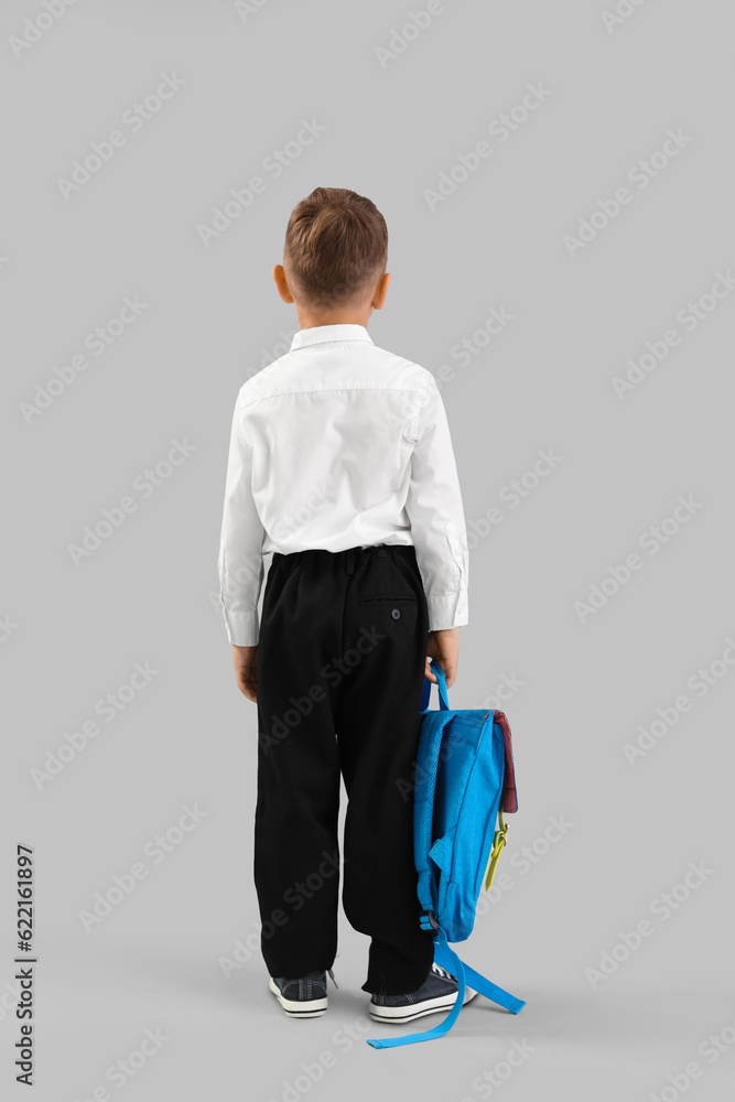 Little schoolboy with blue backpack on grey background, back view