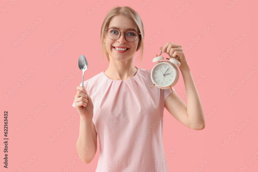 Young woman with spoon and alarm clock on pink background