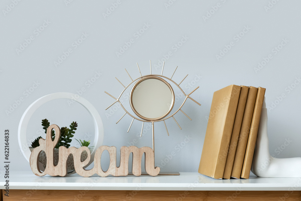 Small mirror, books and florarium on white table near grey wall, closeup