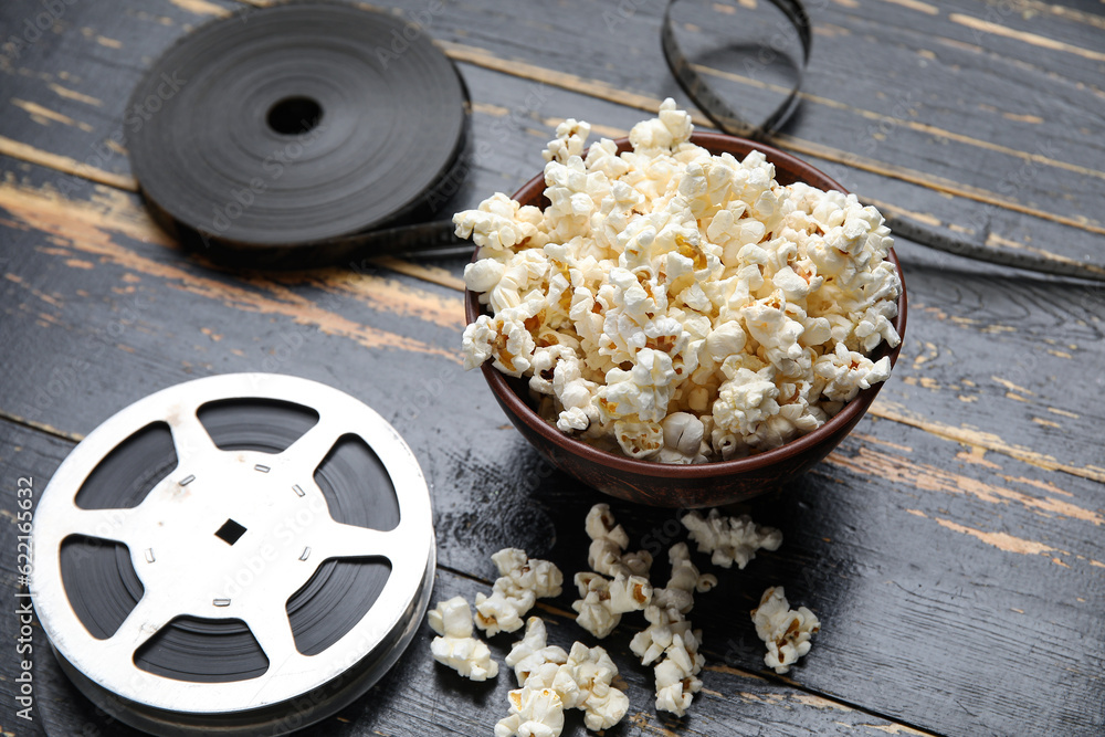 Bowl with tasty popcorn and film reels on black wooden background