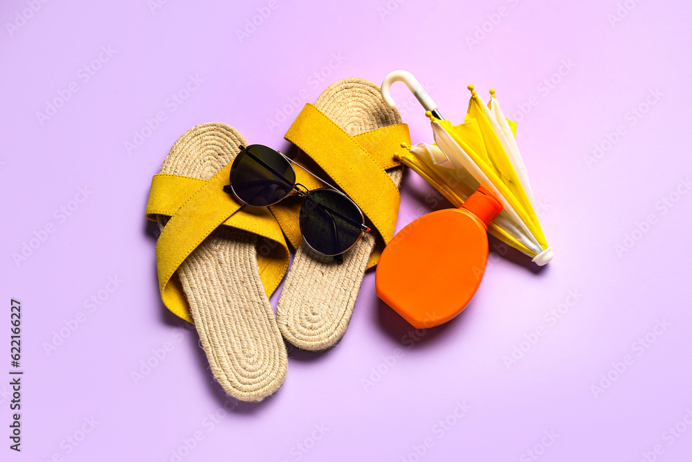 Sunglasses with flip flops, mini umbrella and bottle of sunscreen cream on lilac background