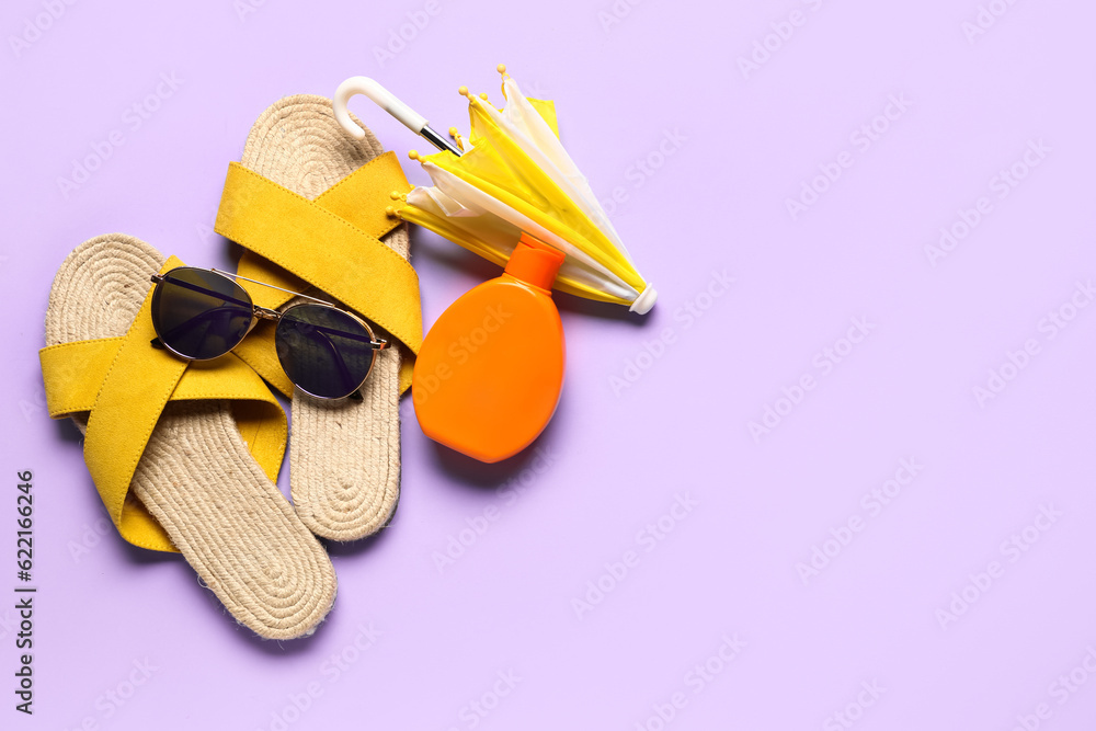 Sunglasses with flip flops, mini umbrella and bottle of sunscreen cream on lilac background