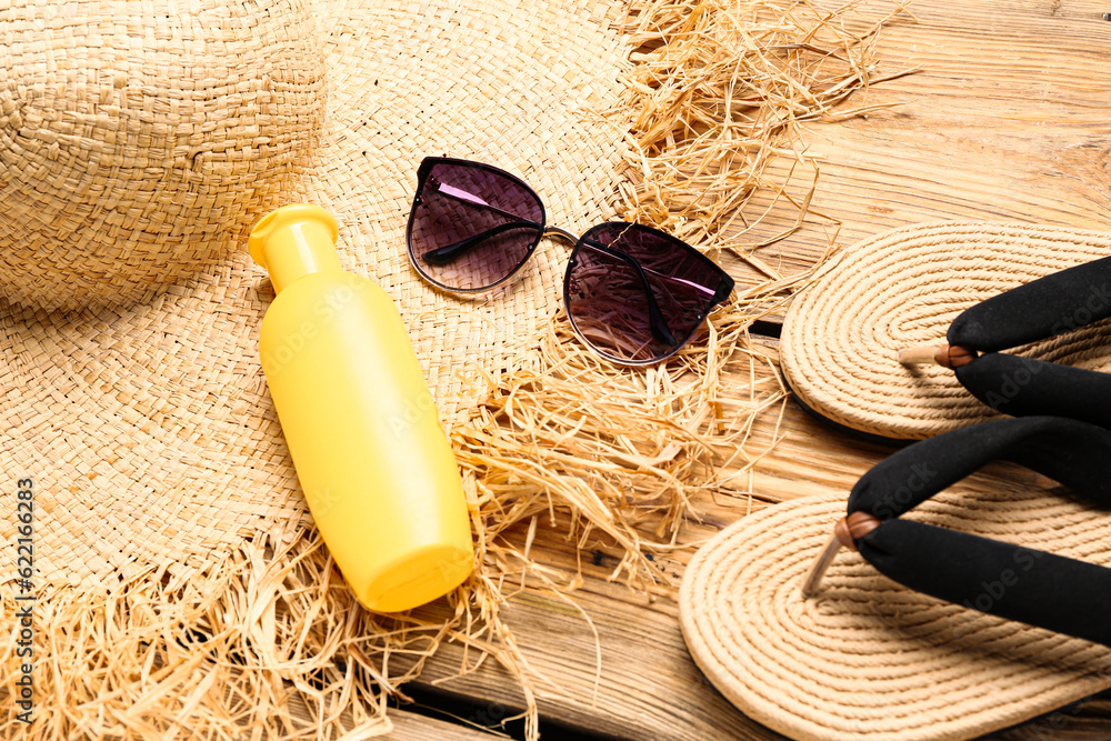 Sunglasses with flip flops, wicker hat and bottle of sunscreen cream on brown wooden background