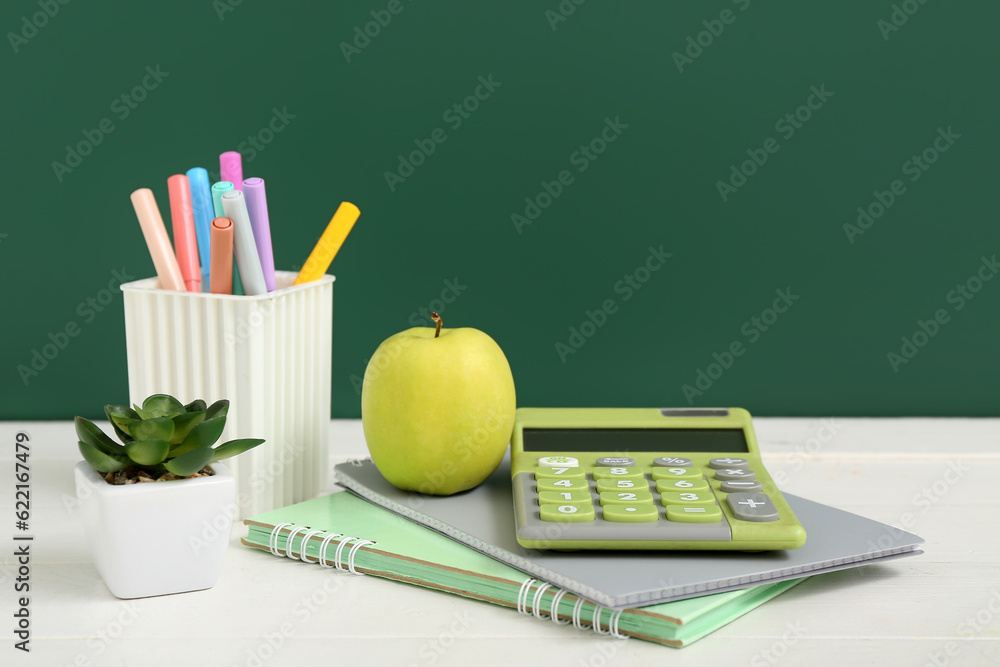 Different stationery with fresh apple and calculator on white table against green chalkboard