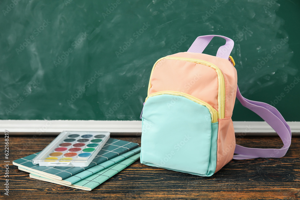 Colorful school backpack with notebooks and watercolors on wooden table near green chalkboard