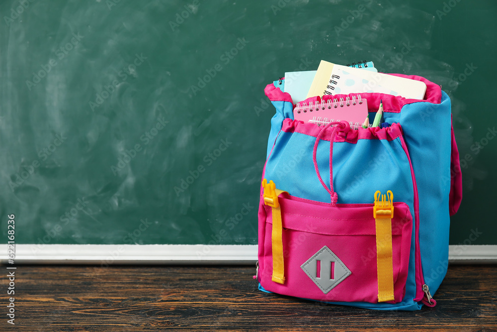 Colorful school backpack with notebooks and pencils on wooden table near green chalkboard