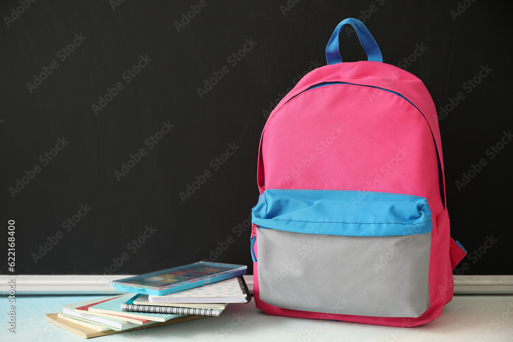 Colorful school backpack with notebooks and watercolors on white table near black chalkboard