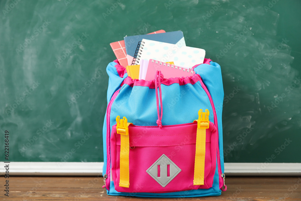 Colorful school backpack with notebooks on wooden table near green chalkboard