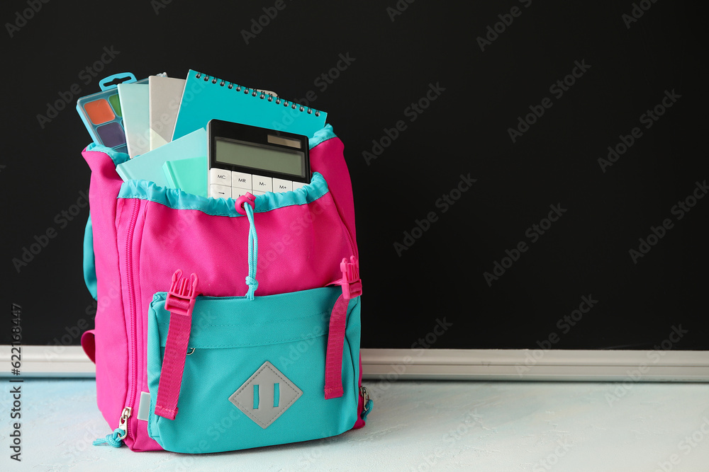 Colorful school backpack with notebooks, calculator and watercolors on white table near black chalkb