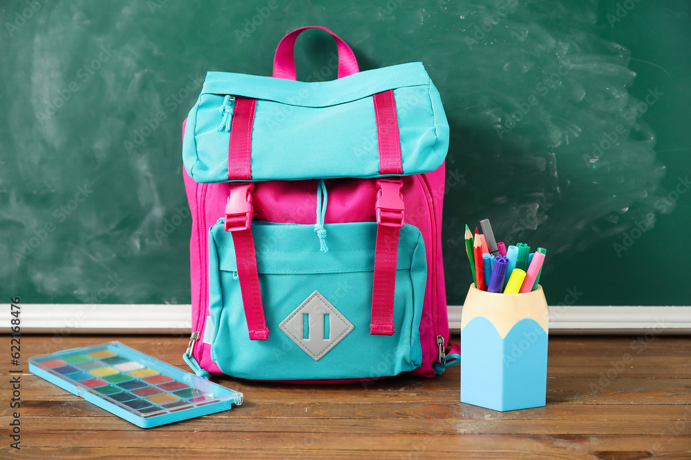 Colorful school backpack with cup of markers and watercolors on wooden table near green chalkboard