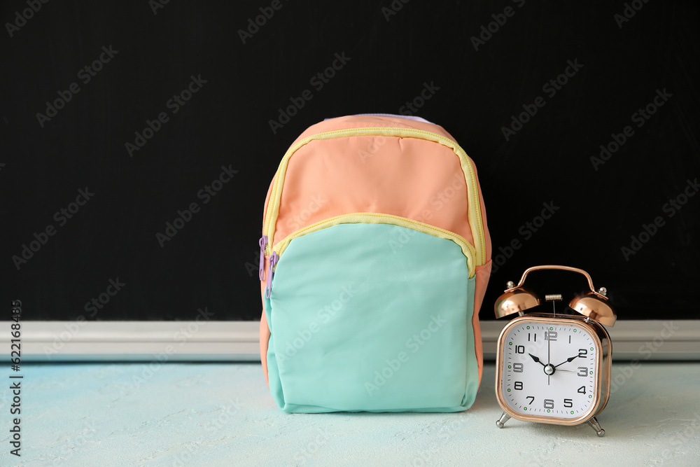 Colorful school backpack with alarm clock on white table near black chalkboard