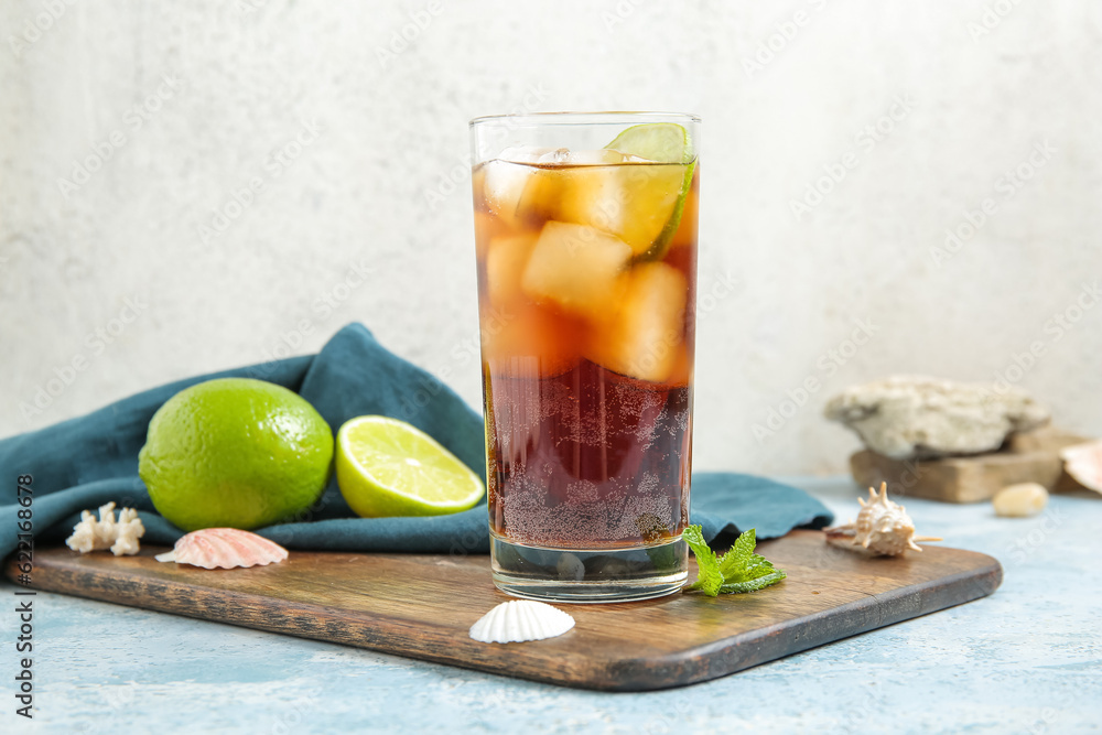 Board with glass of cold Cuba Libre cocktail and seashells on blue table